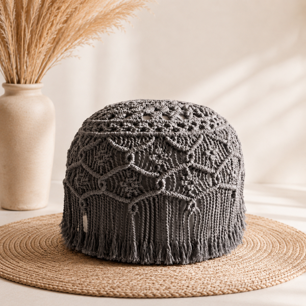 A dark gray handmade macrame pouf with intricate lace-like patterns and fringe, displayed on a woven straw hat stand with a beige ceramic vase of dried grass in the background.