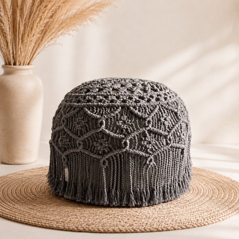 A dark gray handmade macrame pouf with intricate lace-like patterns and fringe, displayed on a woven straw hat stand with a beige ceramic vase of dried grass in the background.