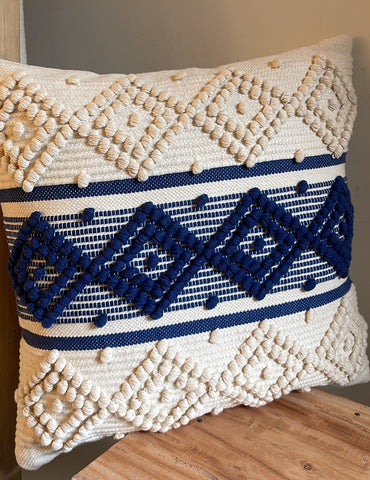 A square woven throw pillow with a cream and navy blue geometric pattern, resting on a wooden table against a neutral wall.
