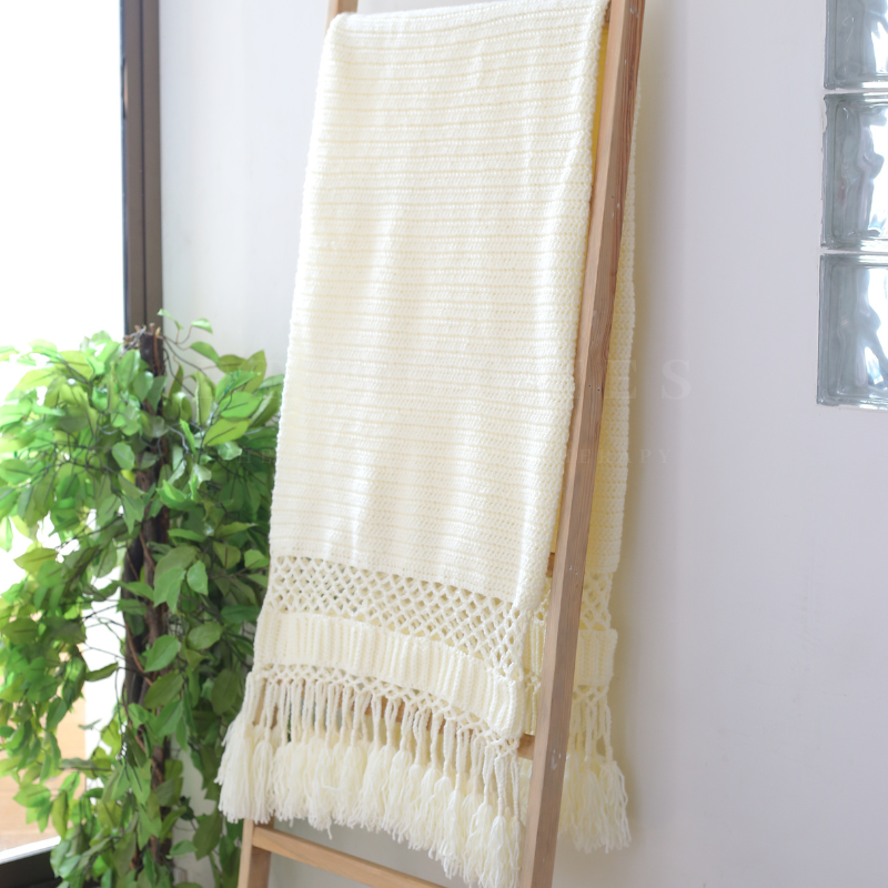 White Woven throw blanket with fringe on a wooden ladder against a white wall with a plant in the background.